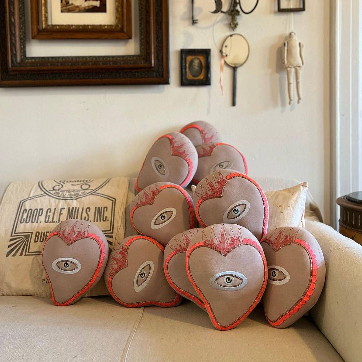 The Skippy Cotton Sacred Heart Pillow—handmade and cotton-filled with stitched eyes and red flame borders—rests on a beige couch in a cozy room adorned with whimsical decor, framed art, a doll, and charming accents.