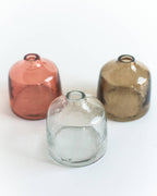 Three small Bella Handblown Hammered Glass Bud Vases: Blush by Creative Women, in pink, brown, and clear, are arranged on a white surface with rounded shapes and narrow openings.