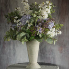 A tall stone vase filled with Ragon House’s Faux White Cherry Blossom Spray and green foliage sits on a round table against a textured gray background.