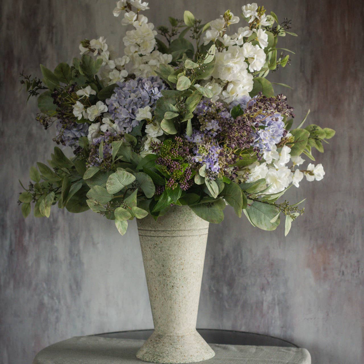 A tall stone vase filled with Ragon House’s Faux White Cherry Blossom Spray and green foliage sits on a round table against a textured gray background.