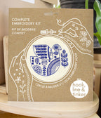 A brown package labeled "Hygge Dove Complete Embroidery Kit" shows blue stitched botanical designs in an embroidery hoop, with the brand "Hook, Line & Tinker Embroidery Kits" at the bottom right.