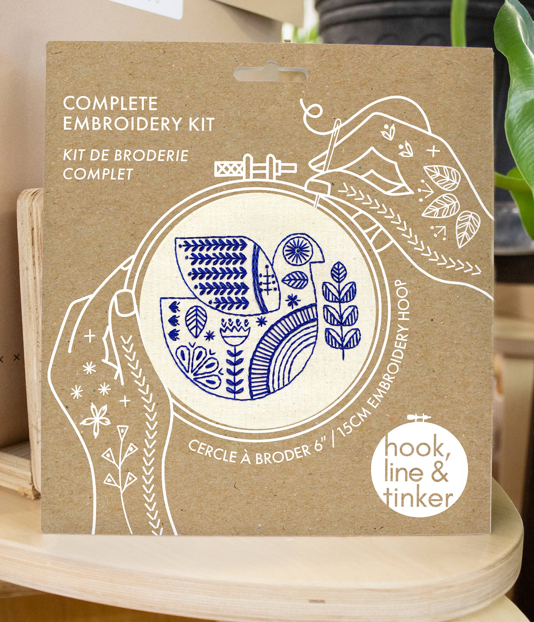 A brown package labeled "Hygge Dove Complete Embroidery Kit" shows blue stitched botanical designs in an embroidery hoop, with the brand "Hook, Line & Tinker Embroidery Kits" at the bottom right.