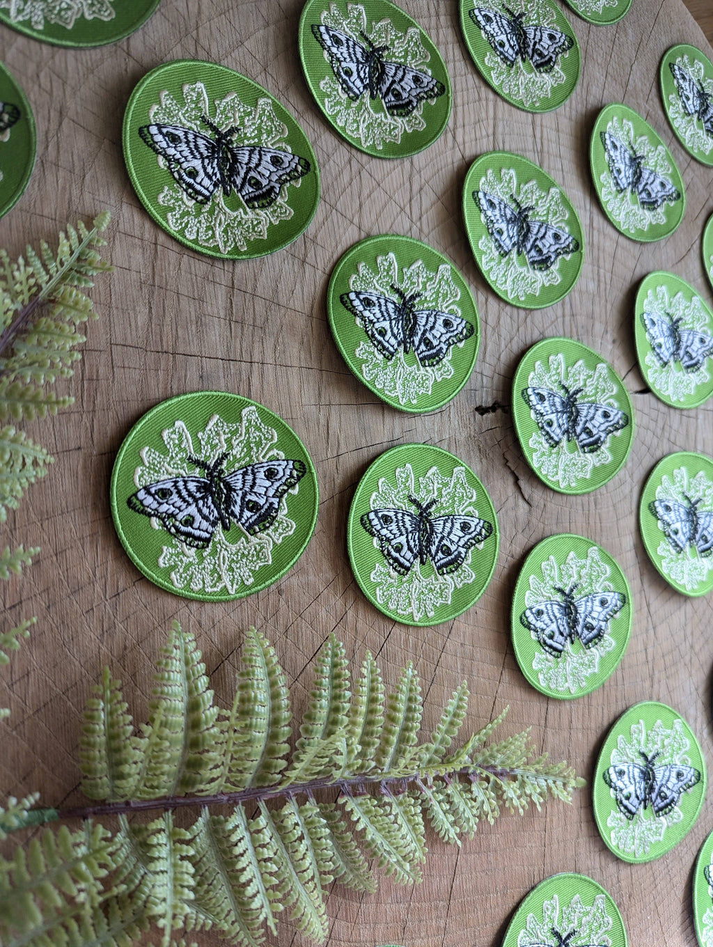 The ilikesara | art + goods iron-on patch features a polyphemus moth with lichen on green, displayed on wood beside an artificial fern.