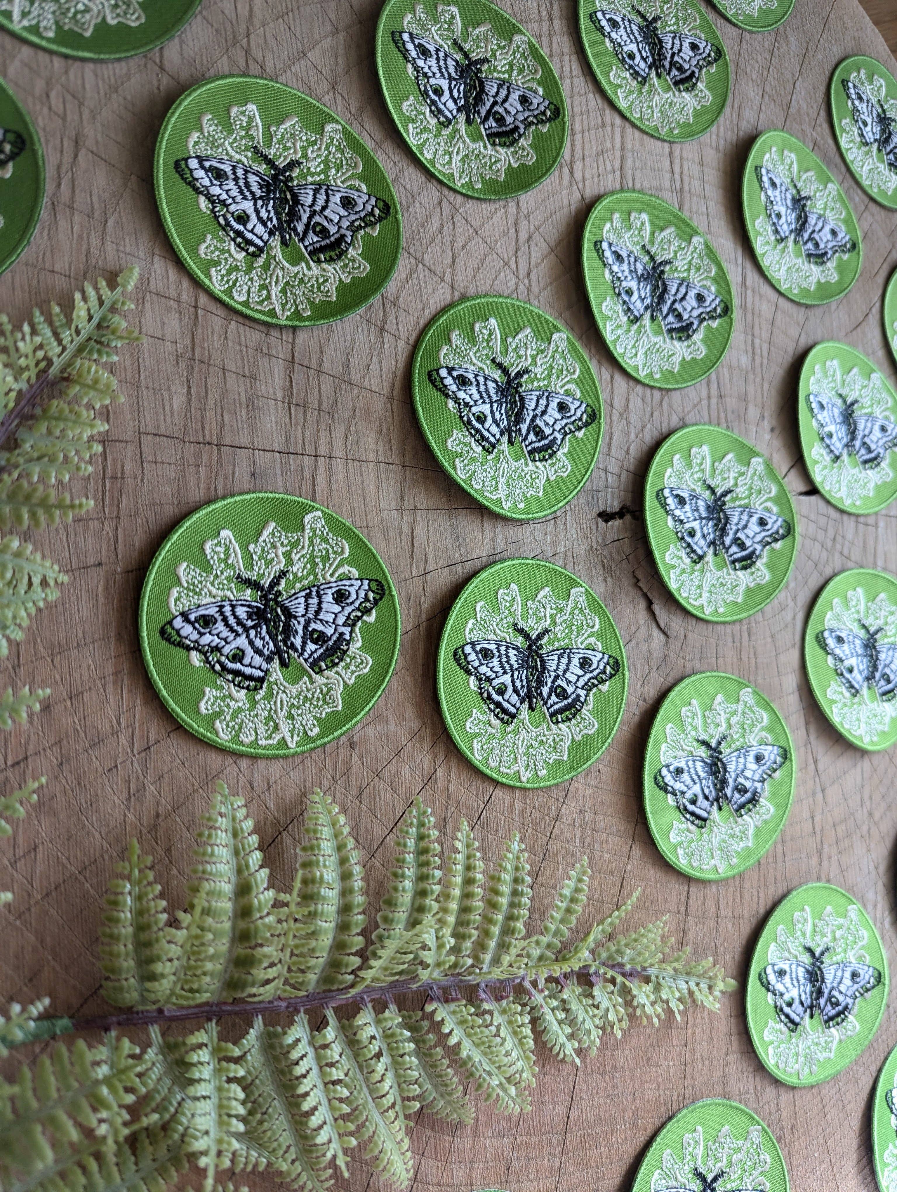 The ilikesara | art + goods iron-on patch features a polyphemus moth with lichen on green, displayed on wood beside an artificial fern.