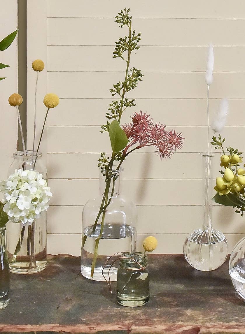 The Clear Medicine Bottle Bud Vase by Jamali Garden displays assorted flowers—white hydrangeas, yellow billy balls, pink fluffy blooms, and green foliage—on a rustic wood surface with a beige paneled wall backdrop.