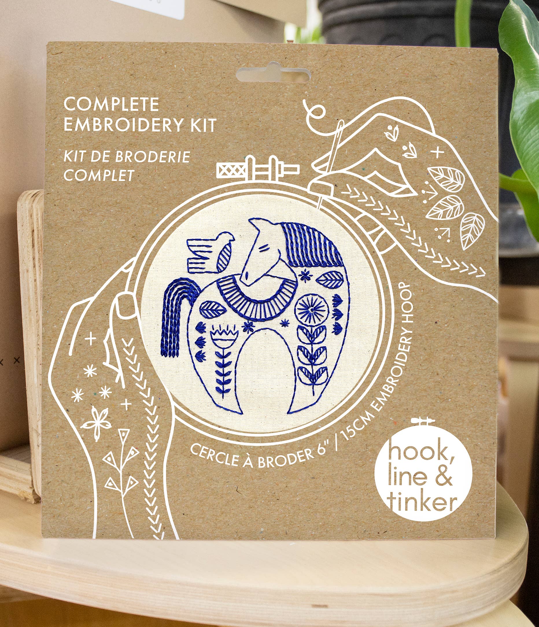The Hygge Horse Complete Embroidery Kit by Hook, Line & Tinker Embroidery Kits features a blue horse design on a hoop with English and French text on brown cardboard; a plant is partly visible on the right.