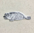 Add the Natural Harp Seal - Gray/White Iron on Applique by Wholesale Applique, an embroidered patch (158470-A) of a cute pup with black eyes on beige fabric—perfect for DIY craft or clothing projects.