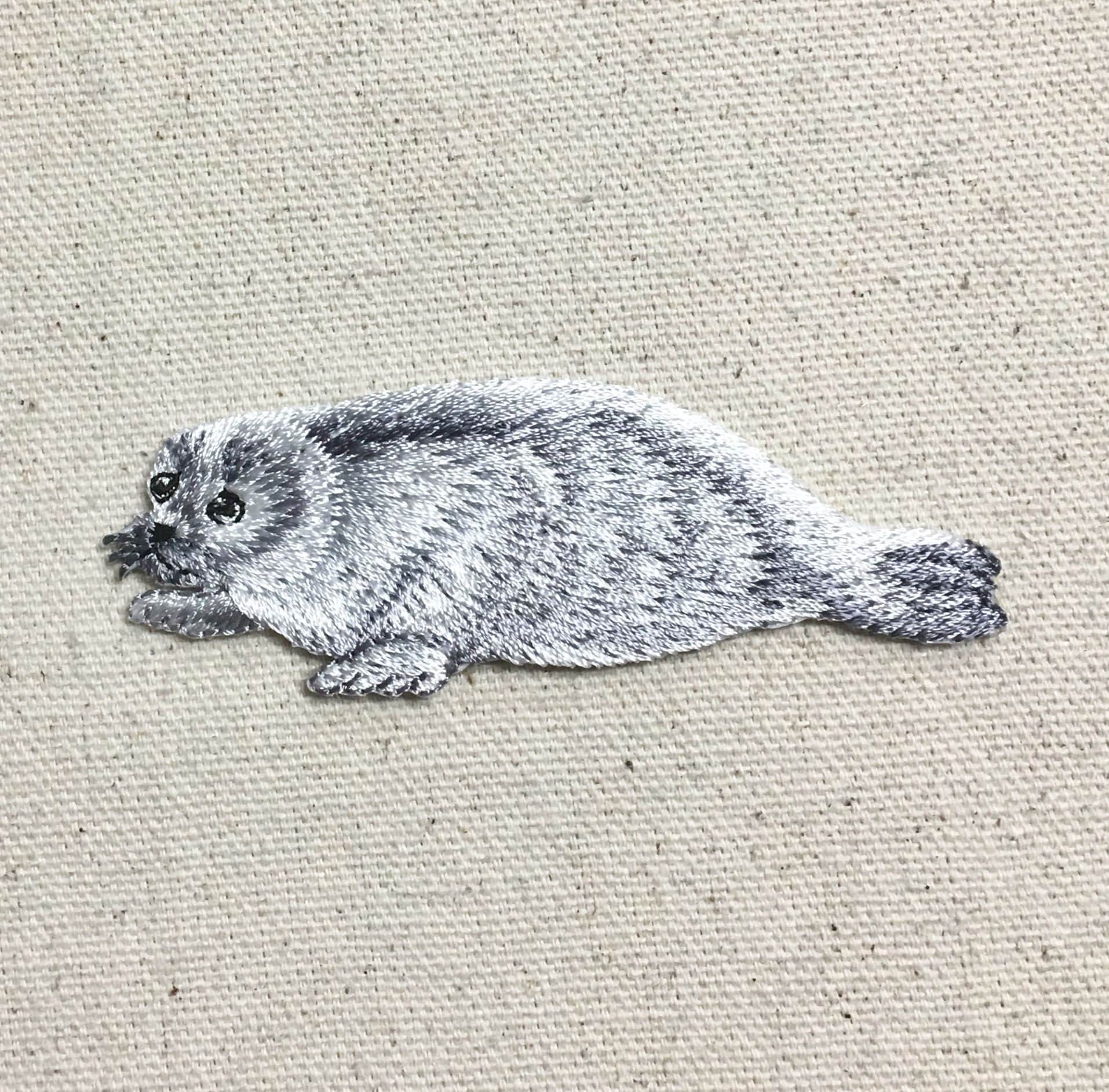 Add the Natural Harp Seal - Gray/White Iron on Applique by Wholesale Applique, an embroidered patch (158470-A) of a cute pup with black eyes on beige fabric—perfect for DIY craft or clothing projects.