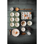 The Danica Heirloom Camellia Bowl Medium 5.75 inch, filled with eggs and nuts, is surrounded by cinnamon sticks, a wooden spoon, milk, and chocolate on a dark wooden surface.