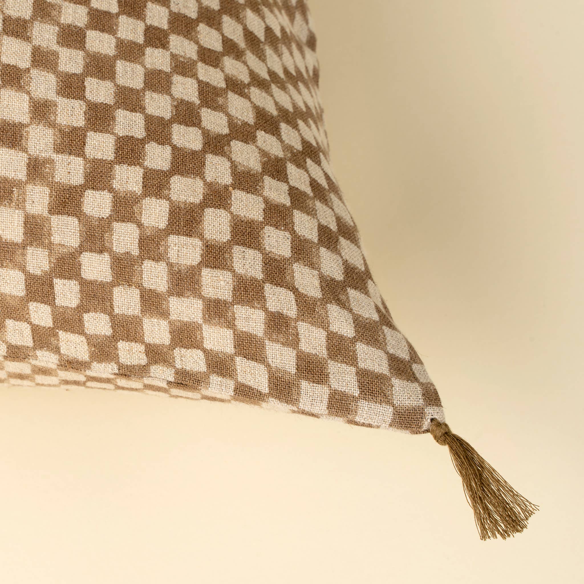 A close-up of the Indaba Trading 16x24 Block Print Check Linen Pillow in brown, featuring a tassel and checkered design, cover with filler, set against a neutral beige background.