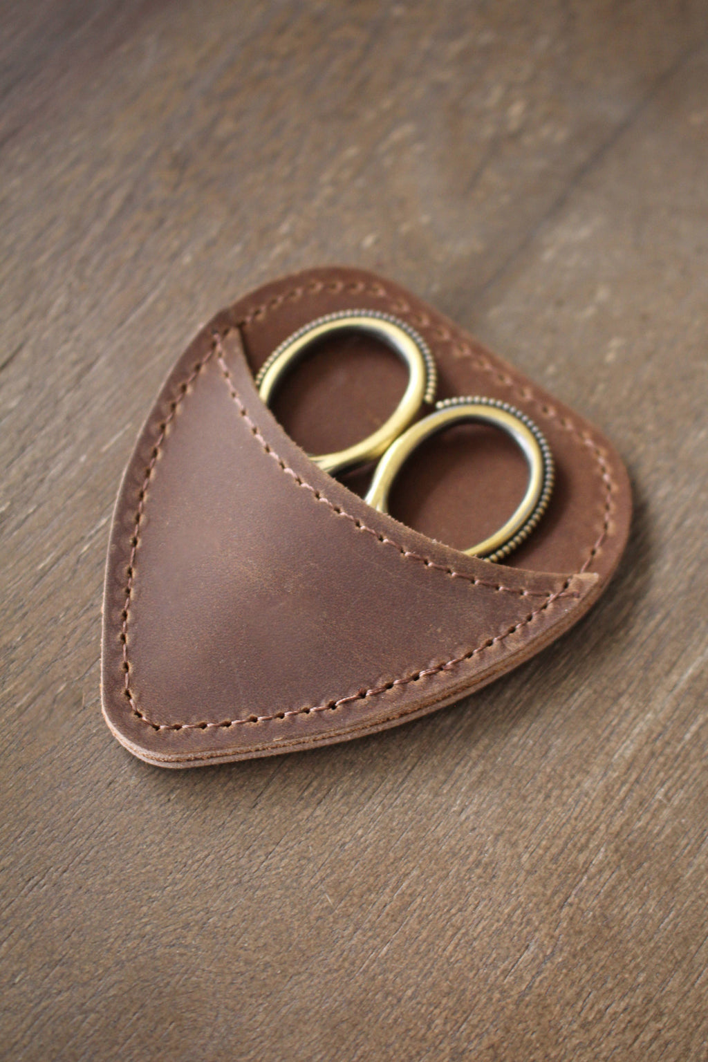 A pair of gold-handled embroidery scissors from NNK Press is partially tucked into a chestnut brown Mini Scissor Keepers leather case, resting on a wooden surface.