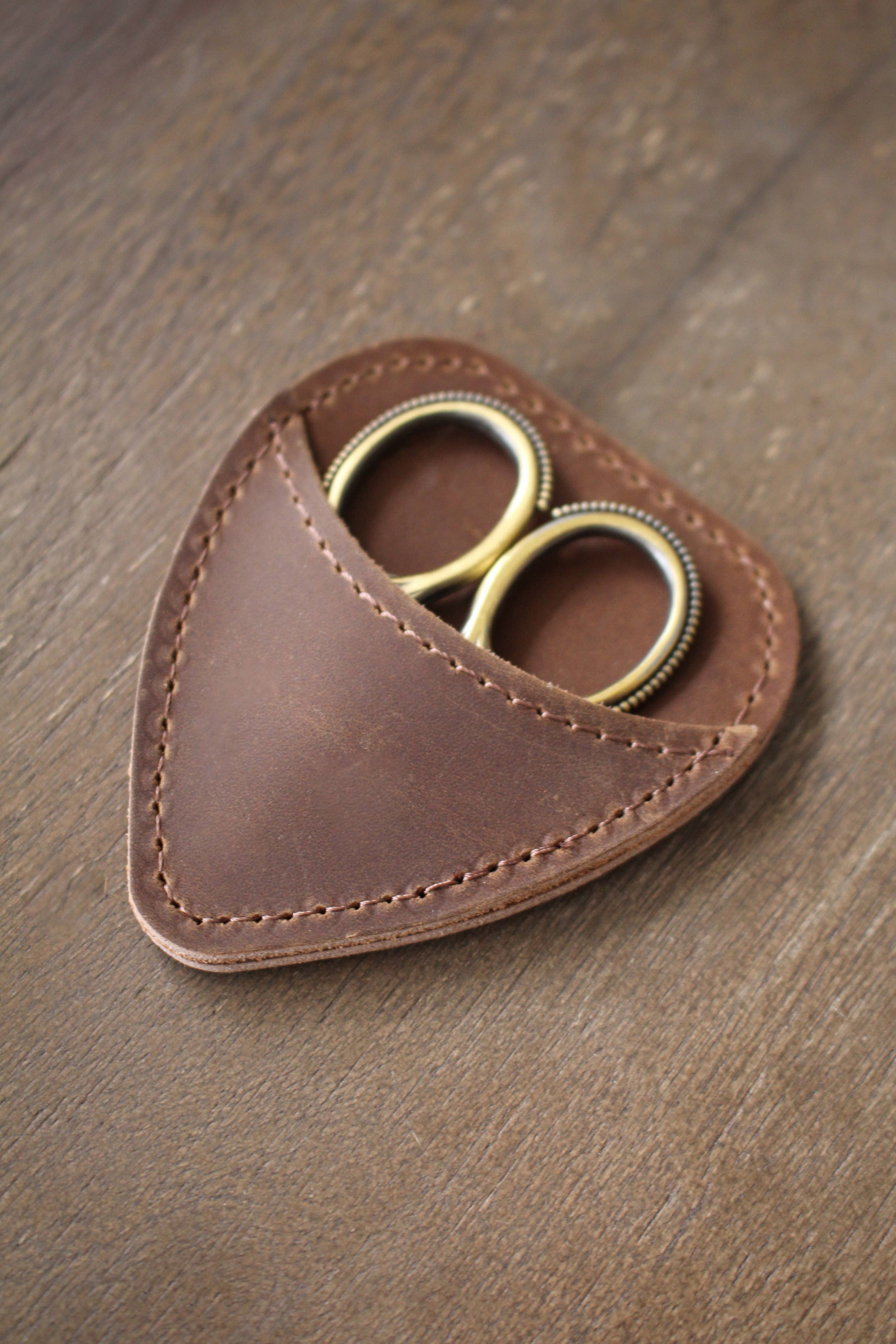 A pair of gold-handled embroidery scissors from NNK Press is partially tucked into a chestnut brown Mini Scissor Keepers leather case, resting on a wooden surface.
