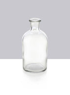 The Jamali Garden Clear Medicine Bottle Bud Vase features a narrow neck and no cap, standing upright on a white surface against a gray background—ideal for stylish home decor.