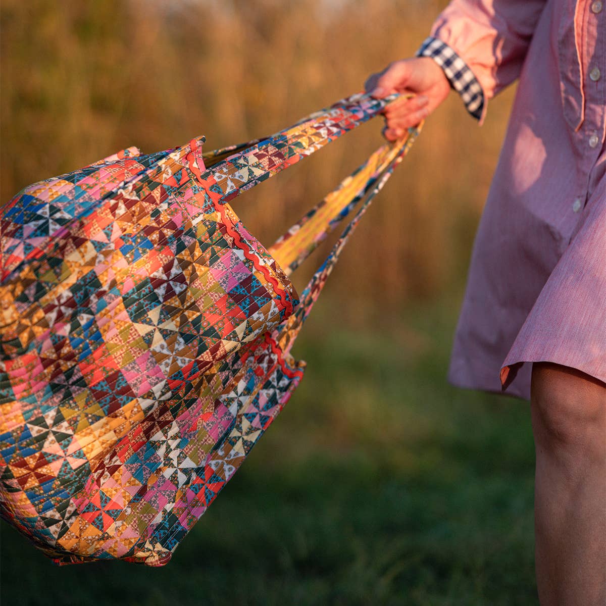 A person in a pink dress holds the Handker Bandanas Calico Quilted Tote outdoors, with green grass and sunlit foliage blurred in the background.