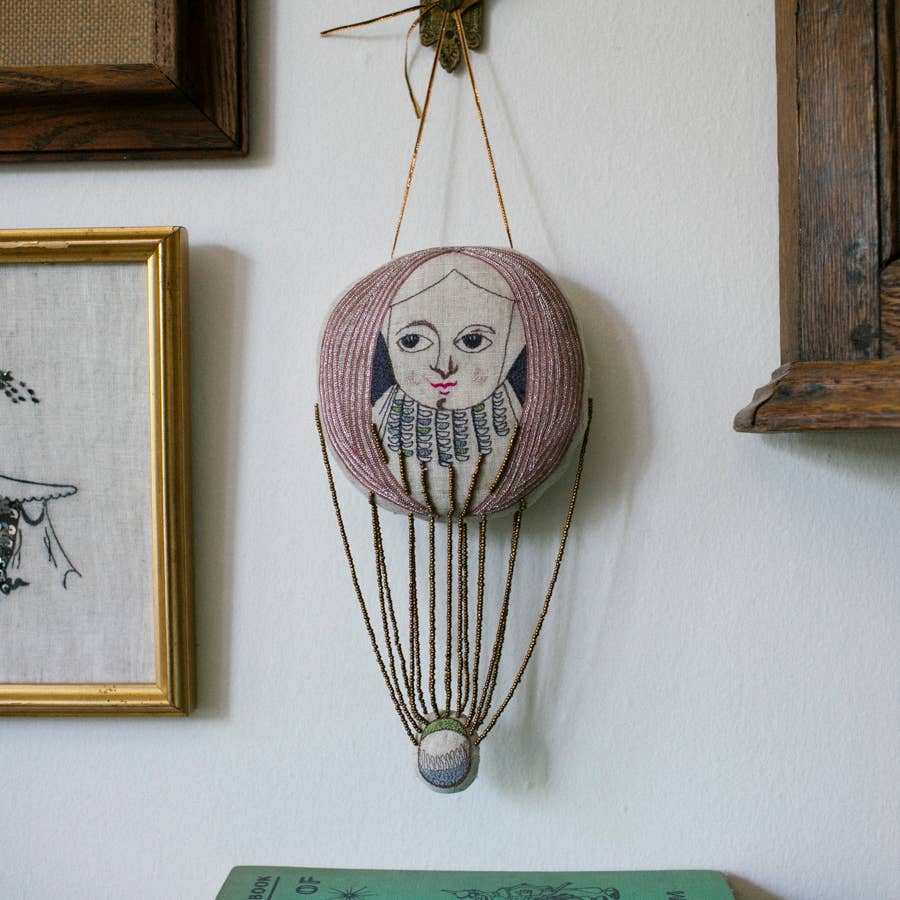The Skippy Cotton Wonderful Wizard / Balloon Oz Wallhanging is a decorative ornament featuring a hand-stitched face, collar, thin ropes, and a fabric basket—perfect as wall art or a unique token for your space.