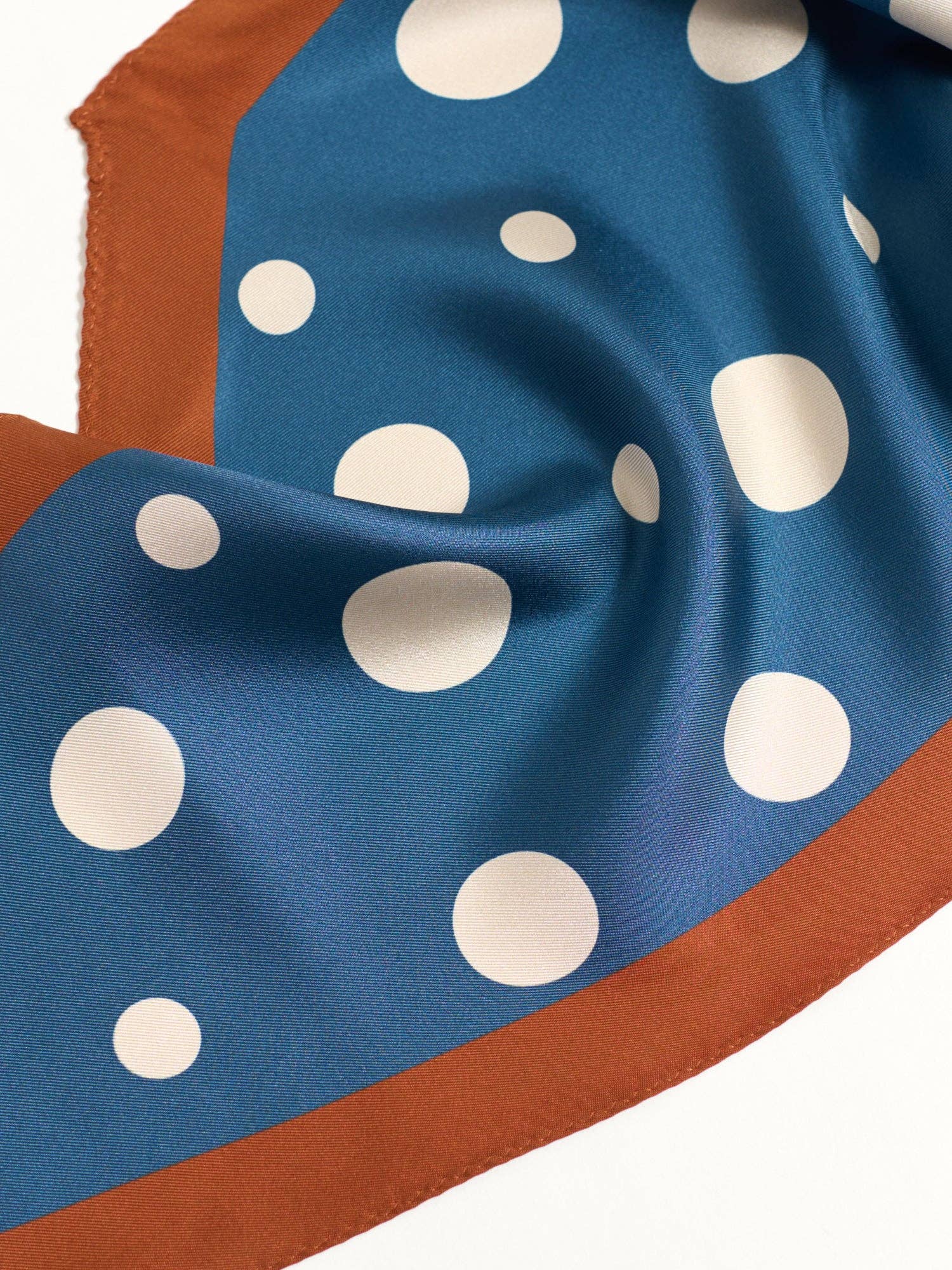 The Silk Polka Dot Diamond Neckerchief - Salted Caramel by Lost Pattern NYC features blue mulberry silk with white polka dots and a rust border, elegantly draped in soft folds on a white background.