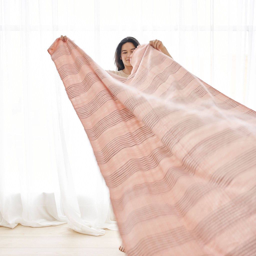 A person holds up and shakes out the Variously Silica Rose Throw in shades of salmon rose and gray stripes in a bright room with sheer white curtains.