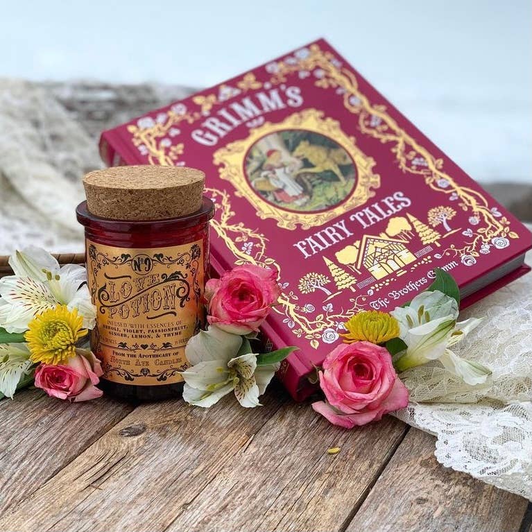 A red and gold Grimm’s Fairy Tales book rests on wood with flowers and lace. Next to it sits North Ave Candles’ The Apothecary / Love Potion / Valentine Candle, its romantic scent enhanced by pink and white blooms.