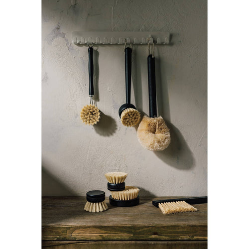 Four natural bristle brushes, including the Danica Heirloom Sisal Palm Dish Brush, hang on a white wall rack above a rustic wood surface with more brushes and a black tray of matches in soft lighting.