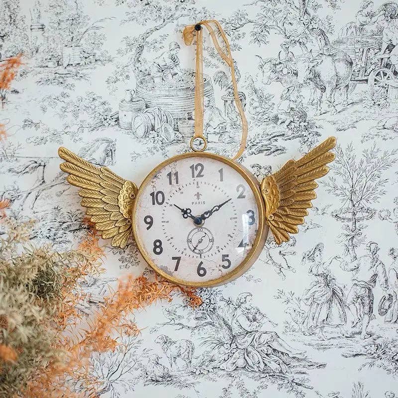 The IRON CLOCK IN GOLD FINISH by Vagabond Vintage, featuring angel wings and ribbon, hangs against black-and-white wallpaper with dried flowers in the lower left corner.