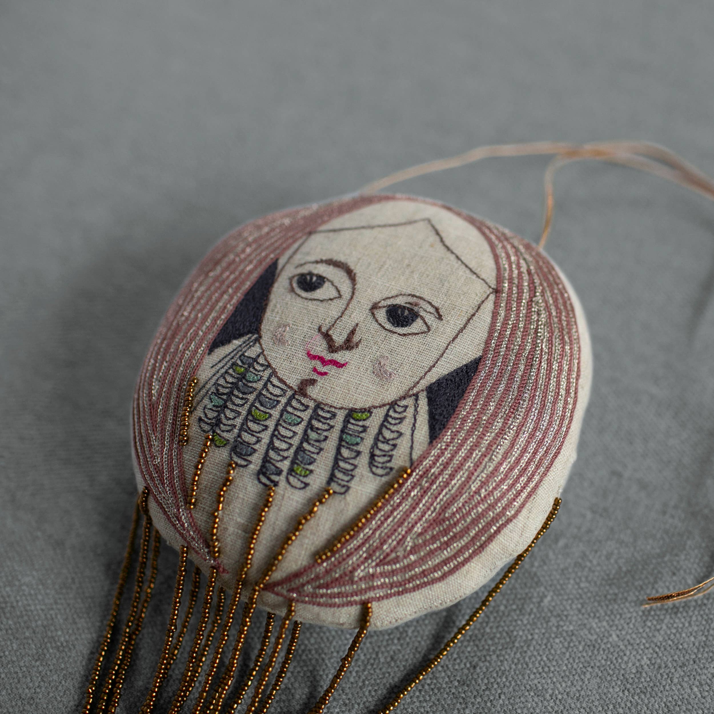 The Skippy Cotton "Wonderful Wizard / Balloon, Oz Wallhanging, Ornament, Token" features a hand-drawn face, long brown hair, stitched patterns, and gold beaded strands—perfect as Wizard of Oz textile art or a unique embroidered wall ornament.