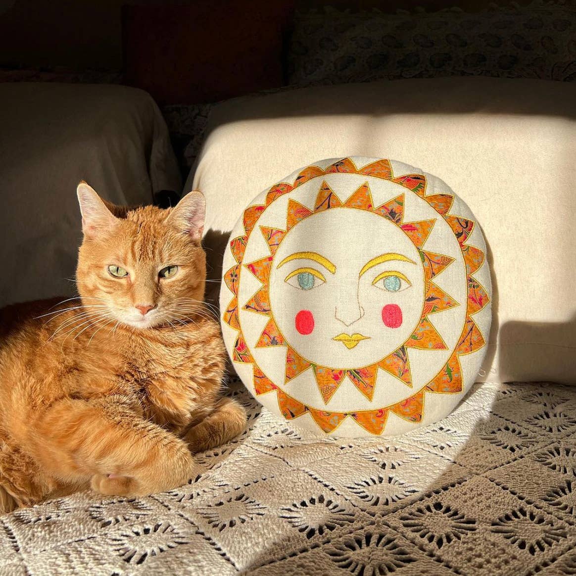 An orange cat relaxes on a crocheted blanket beside the Skippy Cotton Bright Neon Sun Pillow—a handmade, cotton-filled cushion with red cheeks and a yellow nose—both glowing in sunlight.
