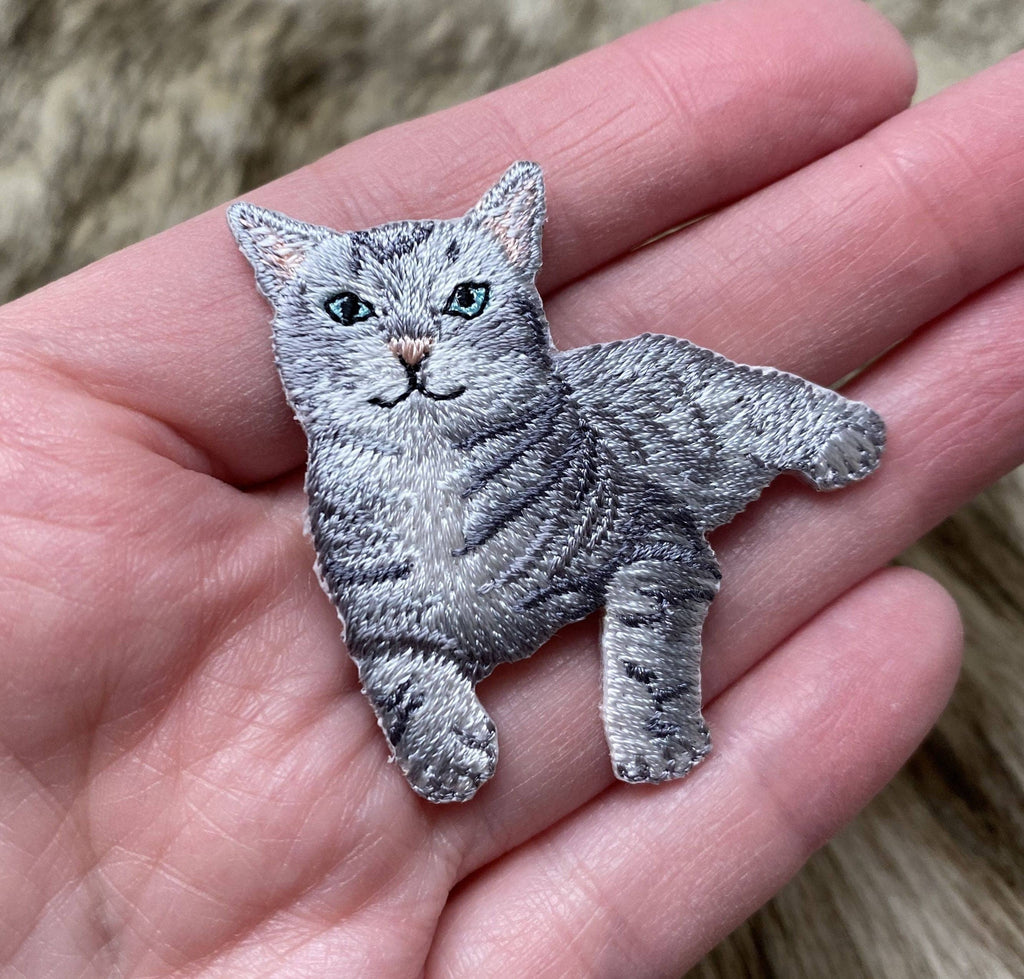 A hand holds a Wholesale Applique Gray Tabby Cat Iron on Patch, featuring green eyes, detailed fur, and black stripes. The blurred background resembles a furry surface.