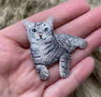 A hand holds a Wholesale Applique Gray Tabby Cat Iron on Patch, featuring green eyes, detailed fur, and black stripes. The blurred background resembles a furry surface.
