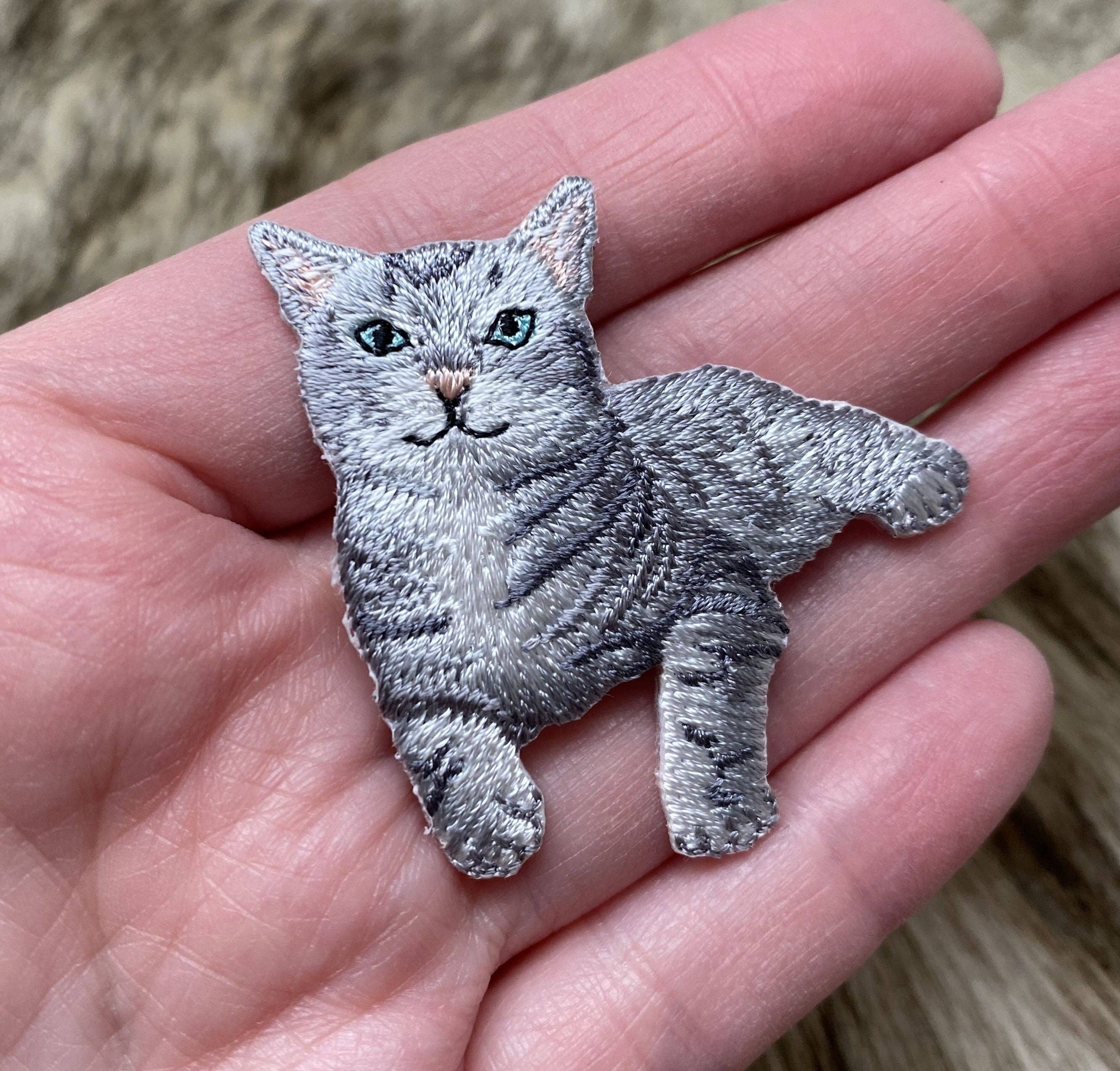 A hand holds a Wholesale Applique Gray Tabby Cat Iron on Patch, featuring green eyes, detailed fur, and black stripes. The blurred background resembles a furry surface.