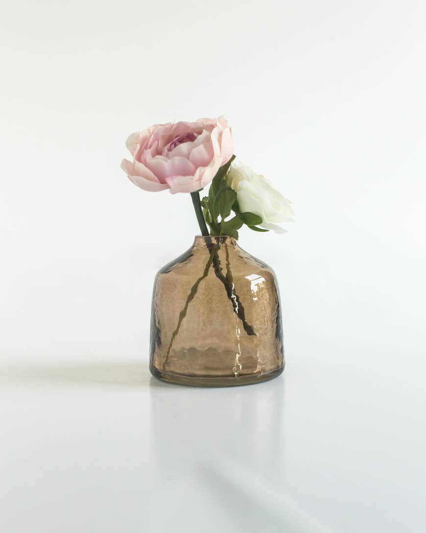 The Bella Handblown Hammered Glass Bud Vase: Blush by Creative Women, a small textured brown vase, holds a pale pink and a white flower against a plain white background.