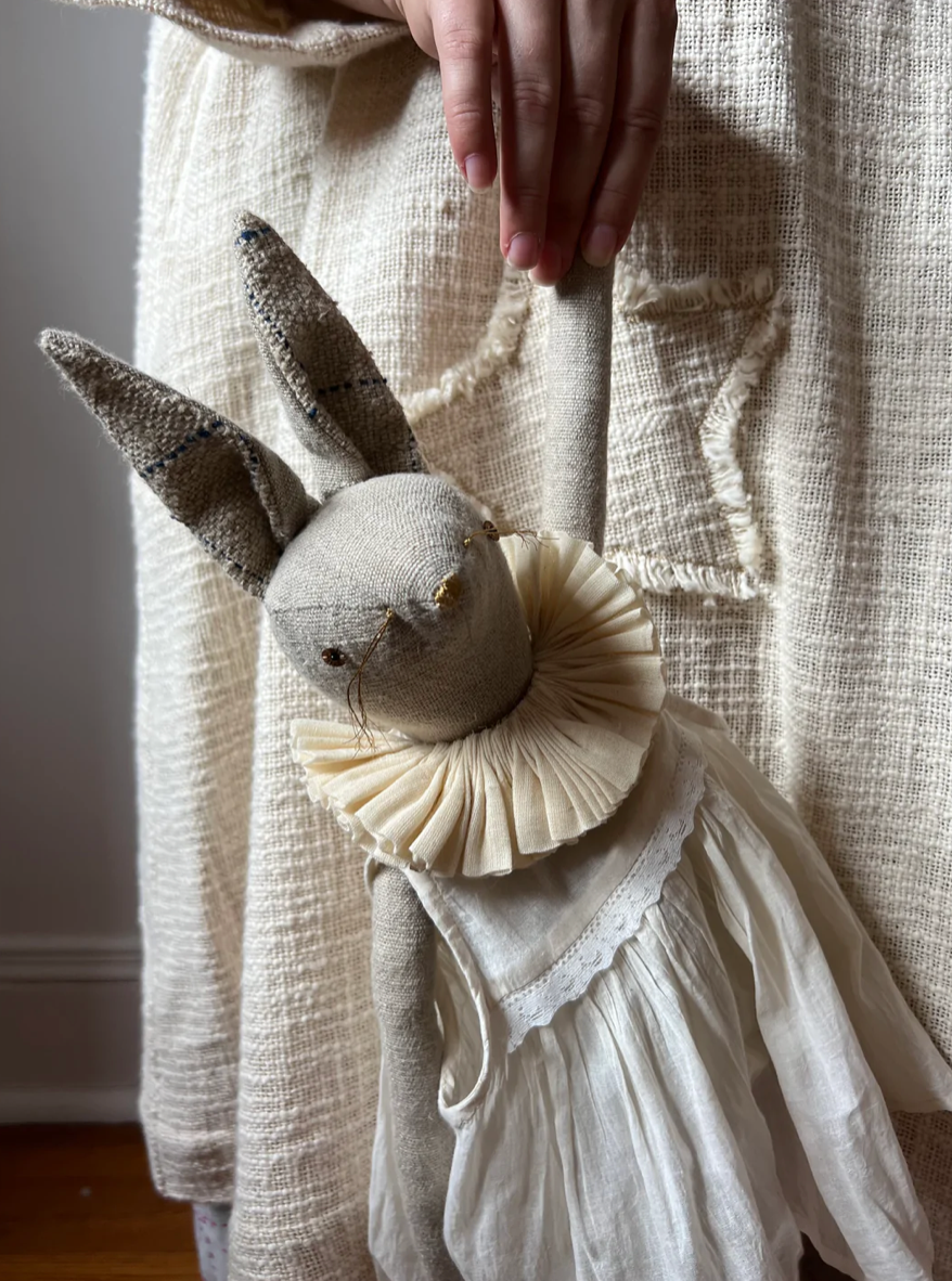 A person wearing a textured, off-white dress holds the Skippy Cotton Ruffle Bun Ragdoll—an art doll cotton-filled and handmade, with a ruffled collar and white dress featuring visible stitching for a gently worn look.