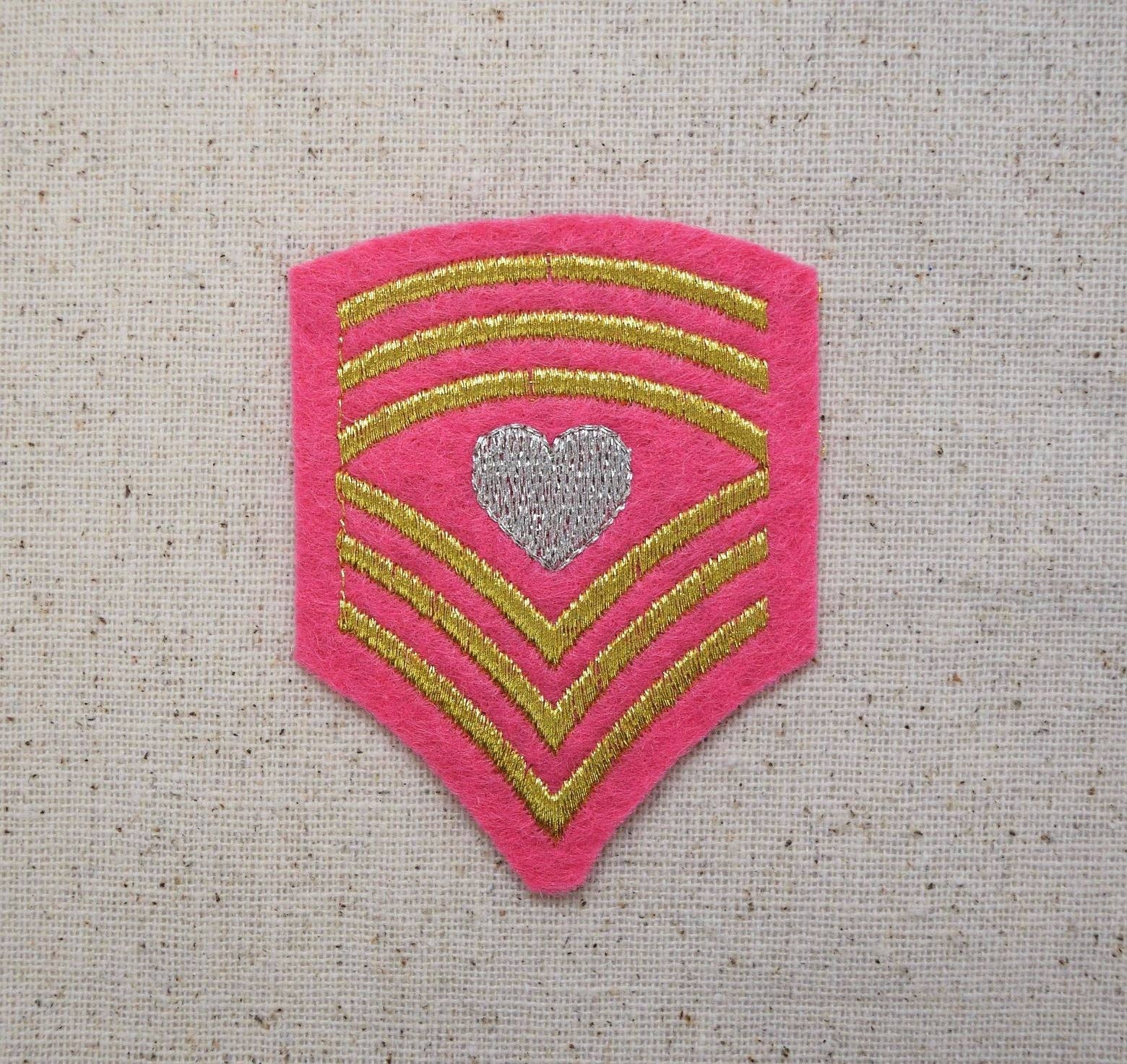 The Wholesale Applique Pink Military Chevron - Shield with Heart embroidered patch features gold stripes and a silver heart, ideal for DIY crafts or as an iron on applique. Style 621022-A is shown on a beige fabric background.