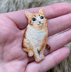 A hand holds the Wholesale Applique Orange Tabby Cat embroidered iron-on patch, featuring a realistic kitten with green eyes sitting upright against a soft, blurred background.