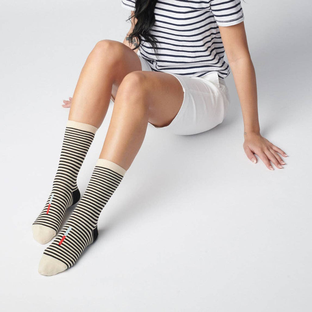 A woman sits on the floor with one hand behind her and legs extended, wearing Cole Black and White Striped Crew Socks by Hooray Sock Co., a black and white striped t-shirt, and white shorts.