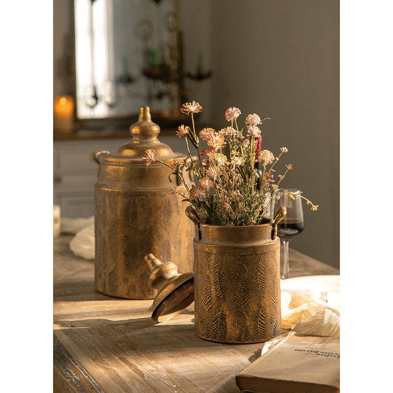 Two Vagabond Vintage Vintage Style Embossed Canister: Large sit on a wooden table; one holds dried flowers with its lid off, the other is closed. Nearby are a glass of red wine and a book in soft lighting.