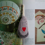 The Skippy Cotton Heart in Hand - Cotton & Lavender filled Ornament, Scented, shaped like a hand with a red heart, sits atop an open book featuring vibrant vintage illustrations about optical toys and art.