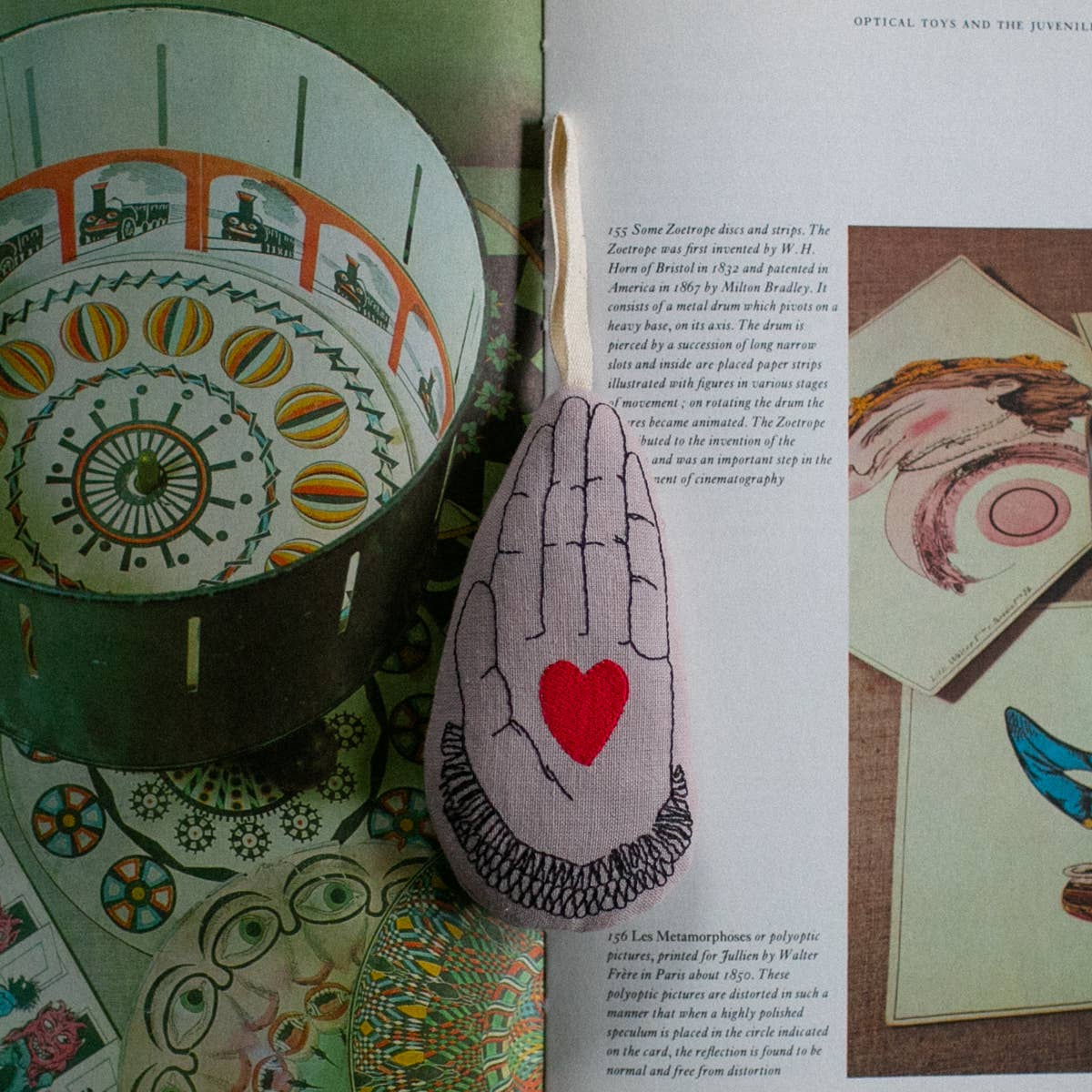 The Skippy Cotton Heart in Hand - Cotton & Lavender filled Ornament, Scented, shaped like a hand with a red heart, sits atop an open book featuring vibrant vintage illustrations about optical toys and art.