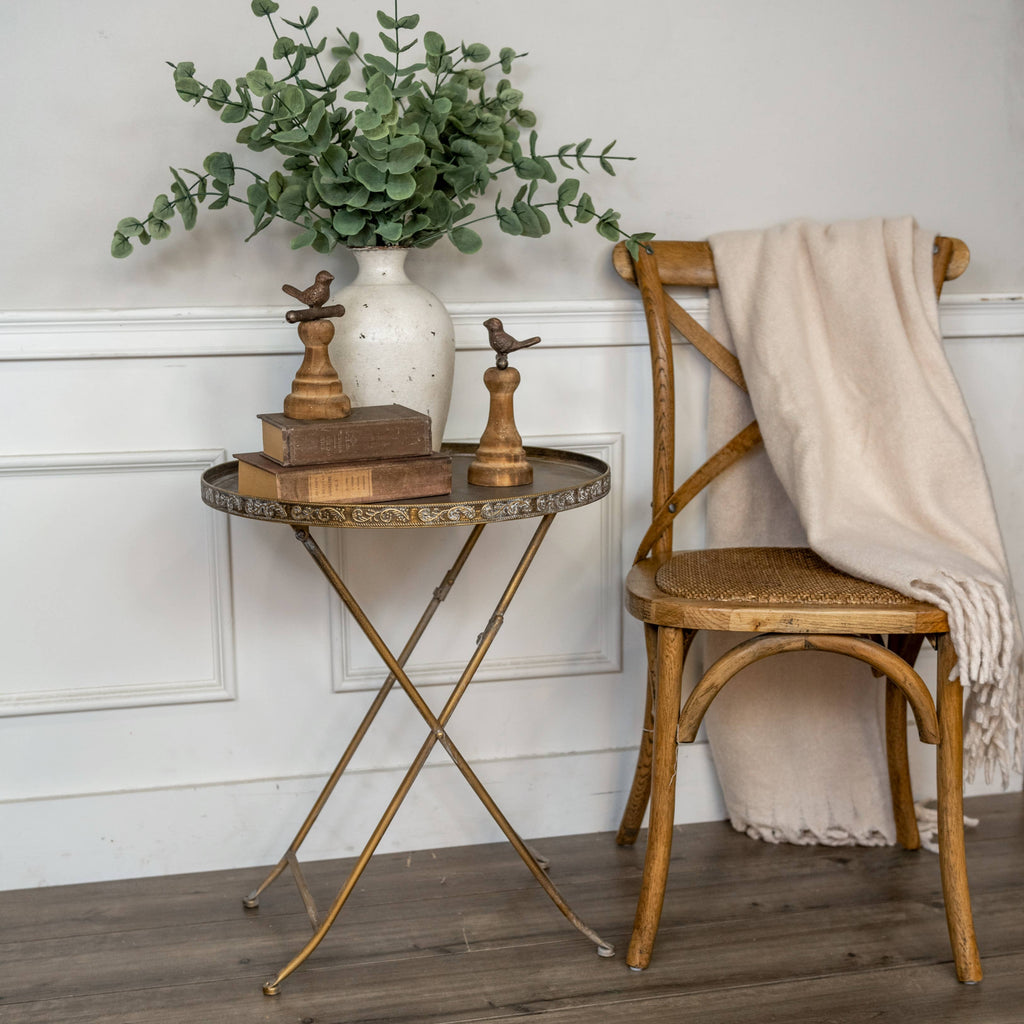 A Ragon House ROUND GOLD FOLDING TRAY TABLE with a potted eucalyptus, books, and bird figurines stands by a wooden chair draped in a beige blanket, set against a white paneled wall and wooden floor.