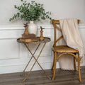 A Ragon House ROUND GOLD FOLDING TRAY TABLE with a potted eucalyptus, books, and bird figurines stands by a wooden chair draped in a beige blanket, set against a white paneled wall and wooden floor.