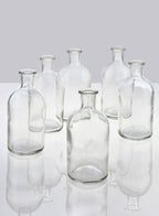 Six empty Jamali Garden Clear Medicine Bottle Bud Vases are arranged on a reflective white surface with a plain light gray background. Similar in size and shape, each vase casts a faint reflection—ideal for minimalist home decor.