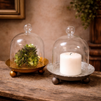 The Cloche Candle Holder with Metal Plate: Gold by Vagabond Vintage displays a green succulent and white pillar candle on a rustic wooden surface, adding charm as elegant home decor. Blurred background accents complete the look.