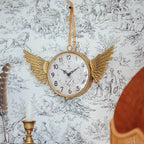 The IRON CLOCK IN GOLD FINISH by Vagabond Vintage hangs on patterned wallpaper at 10:12, radiating whimsy with angel wing details. A nearby candle and feather add charm.