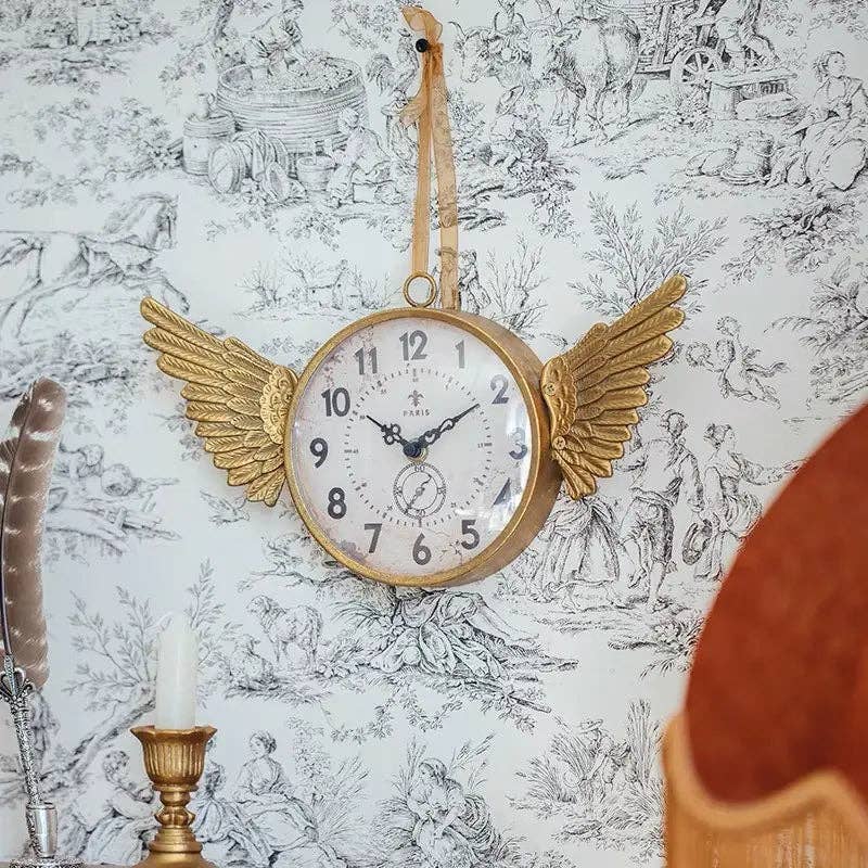 The IRON CLOCK IN GOLD FINISH by Vagabond Vintage hangs on patterned wallpaper at 10:12, radiating whimsy with angel wing details. A nearby candle and feather add charm.