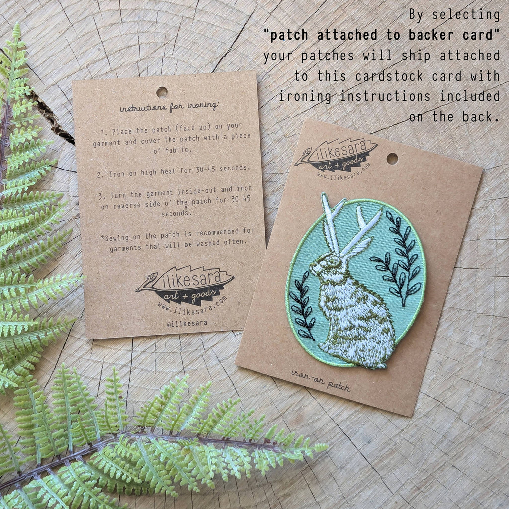 The “Iron-on patch | snail with fern - 2.5\" round teal” by ilikesara | art + goods shows a teal embroidered snail and fern. No backer card or packaging is included. The patch rests directly on a wooden surface.