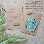 The “Iron-on patch | snail with fern - 2.5\" round teal” by ilikesara | art + goods shows a teal embroidered snail and fern. No backer card or packaging is included. The patch rests directly on a wooden surface.