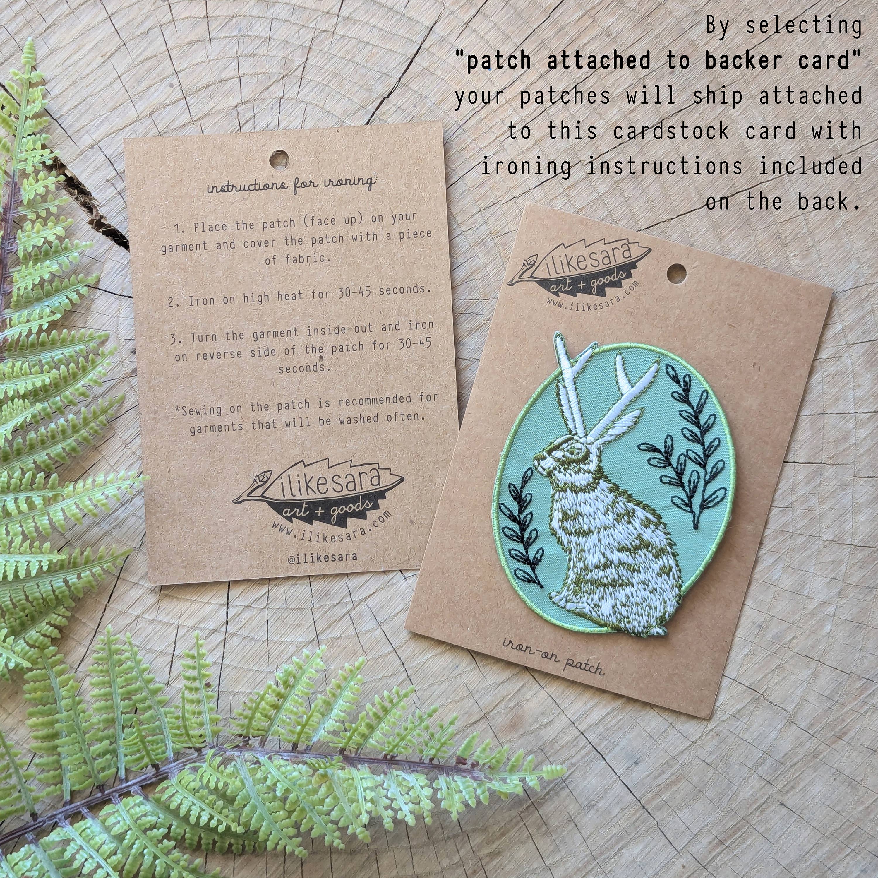 The “Iron-on patch | snail with fern - 2.5\" round teal” by ilikesara | art + goods shows a teal embroidered snail and fern. No backer card or packaging is included. The patch rests directly on a wooden surface.