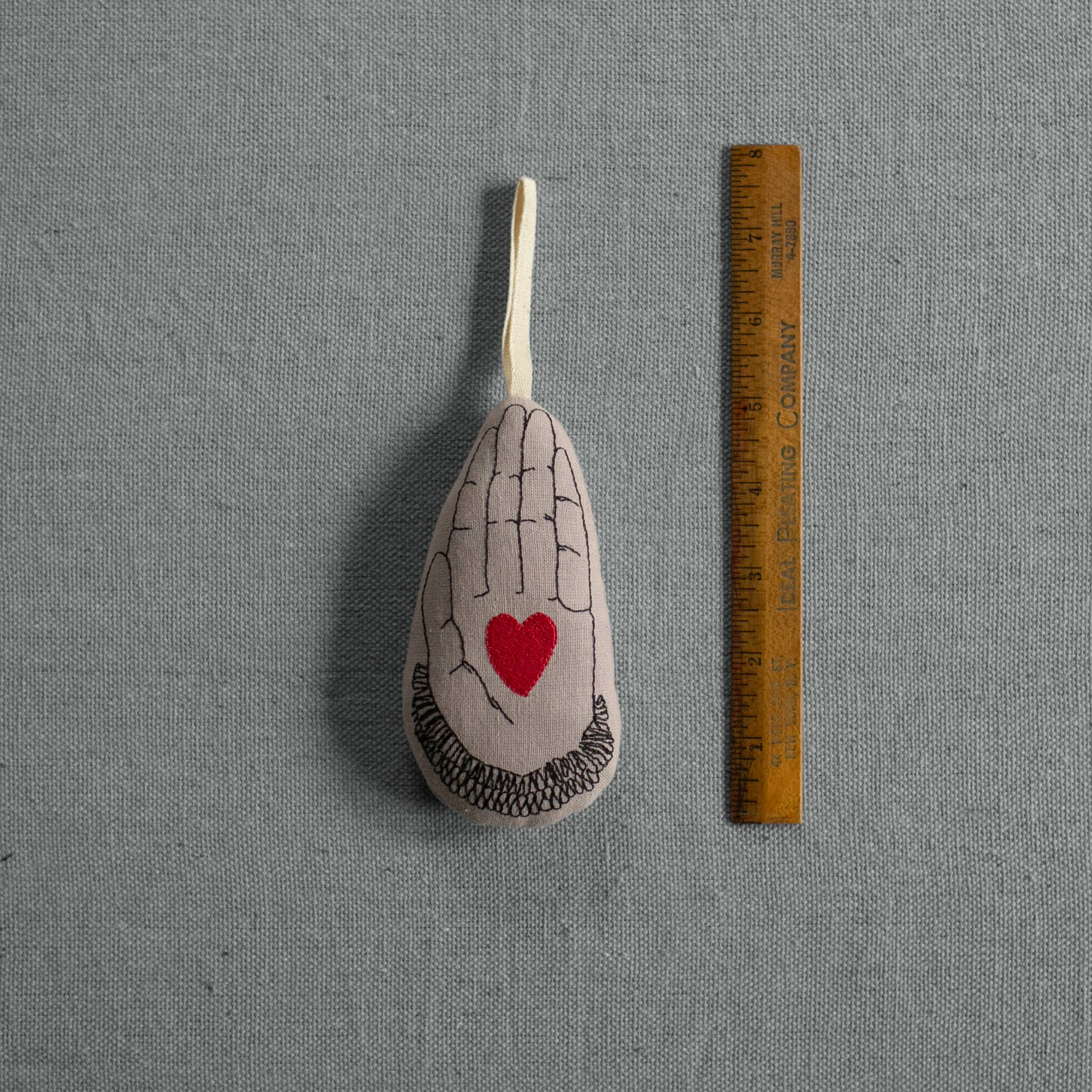 The Skippy Cotton "Heart in Hand" ornament is a small, organic cotton sachet filled with lavender, featuring a printed hand with a red heart. It includes a hanging loop and is shown beside a wooden ruler on a gray textured surface.
