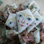 The Skippy Cotton Flying Kite is a diamond-shaped ornament with embroidered faces, red cheeks, and blue eyes, crafted from organic cotton, filled with lavender, and finished with patterned ribbons—perfect as a scented gift topper.