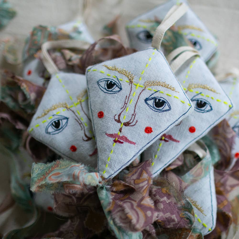 The Skippy Cotton Flying Kite is a diamond-shaped ornament with embroidered faces, red cheeks, and blue eyes, crafted from organic cotton, filled with lavender, and finished with patterned ribbons—perfect as a scented gift topper.
