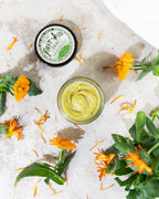 An open jar of Roots And Leaves First Aid Salve rests on a light surface, surrounded by vibrant calendula flowers and petals, with the product’s labeled lid nearby.